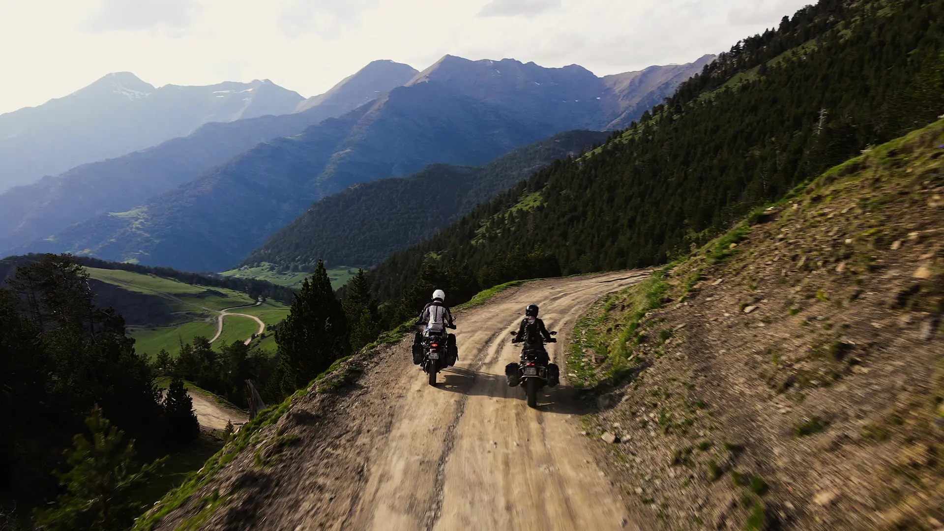 Deux motos de dos sur une piste en balcon surplombant une vallée très encaissée et verdoyante en Andorre.